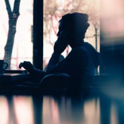 Silhouetted figure using phone and laptop at a cafe.